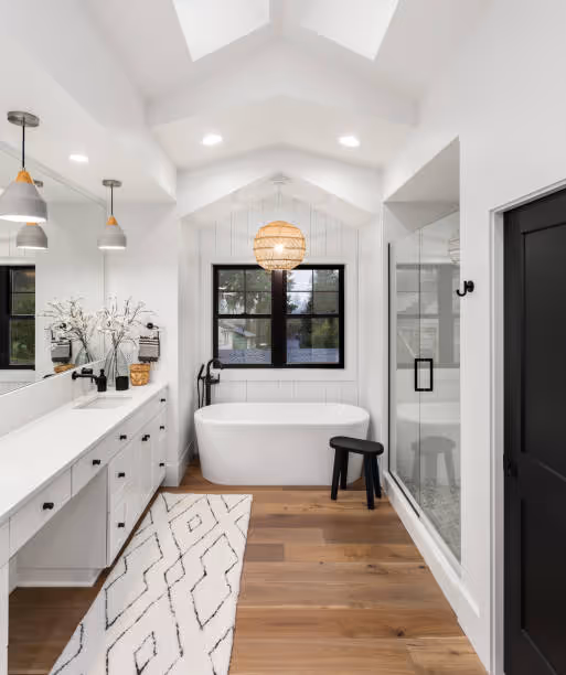 Bright modern bathroom with a freestanding white tub, black-framed window, wooden stool, glass-enclosed shower, and white vanity with pendant lights.