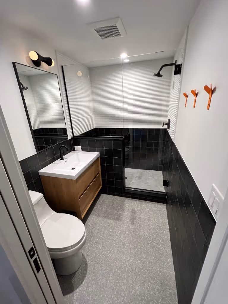 Modern bathroom with black tiled half walls, wood vanity with white sink, white toilet, glass shower enclosure, and three orange wall hooks.