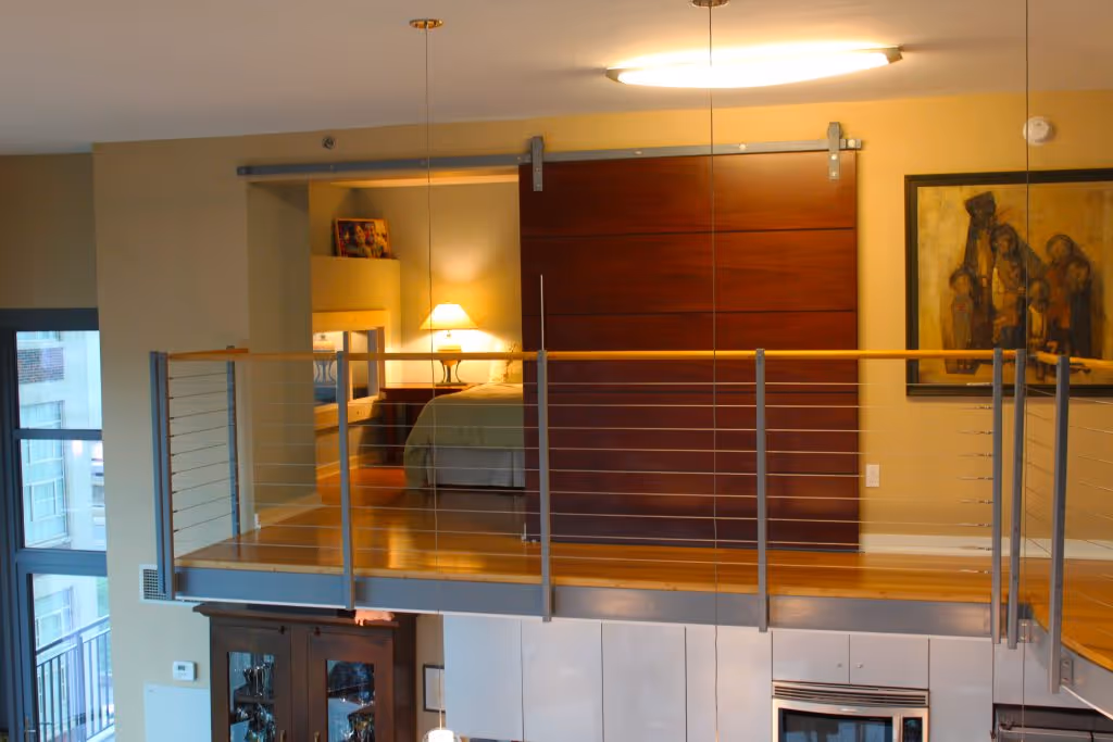 Modern loft bedroom with glass railing, sliding wooden door, bedside lamp, and framed painting on wall.