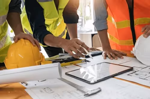 Construction workers in safety vests reviewing blueprints and using a tablet at a worksite.