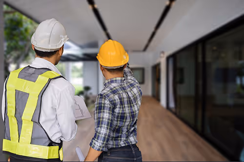 Two construction workers wearing helmets and safety vests discussing plans in a modern building hallway.