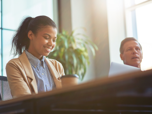 Smiling businesswoman working at a desk with a coffee cup, with a man in the background.