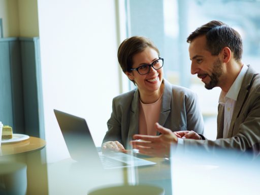 Two business professionals smiling and discussing work on a laptop in a bright office setting.
