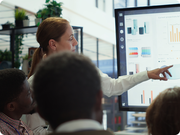 Woman pointing at a large screen displaying financial charts while colleagues watch attentively.