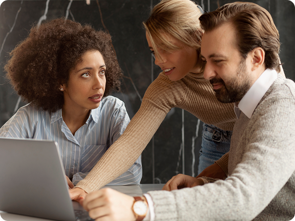 Three colleagues gathered around a laptop, engaged in a discussion with one woman pointing at the screen.