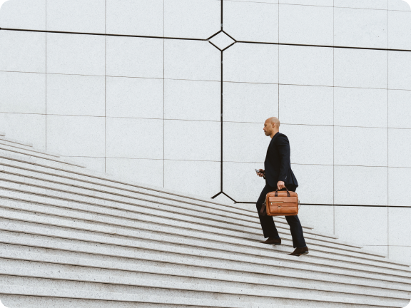 Man in a black suit carrying a brown briefcase walking up wide stone stairs with a large tiled wall in the background.