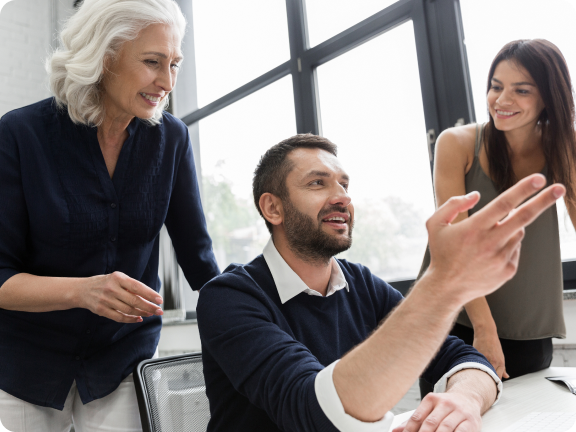 Three colleagues gathered around a desk in an office, a man seated gestures while two women stand and smile attentively.