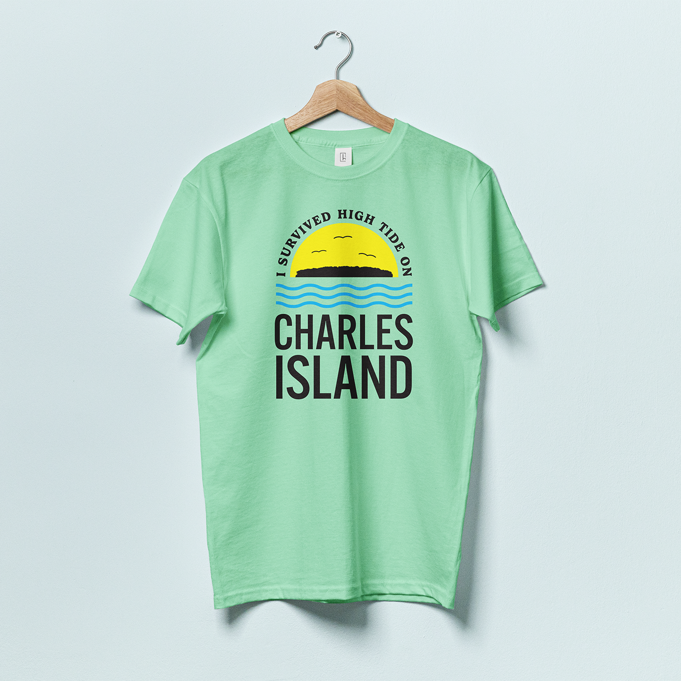 A green t-shirt with a silhouette of Charles Island on top of a yellow semi circle, with blue wavy lines beneath and the words "I survived high tide on Charles Island"