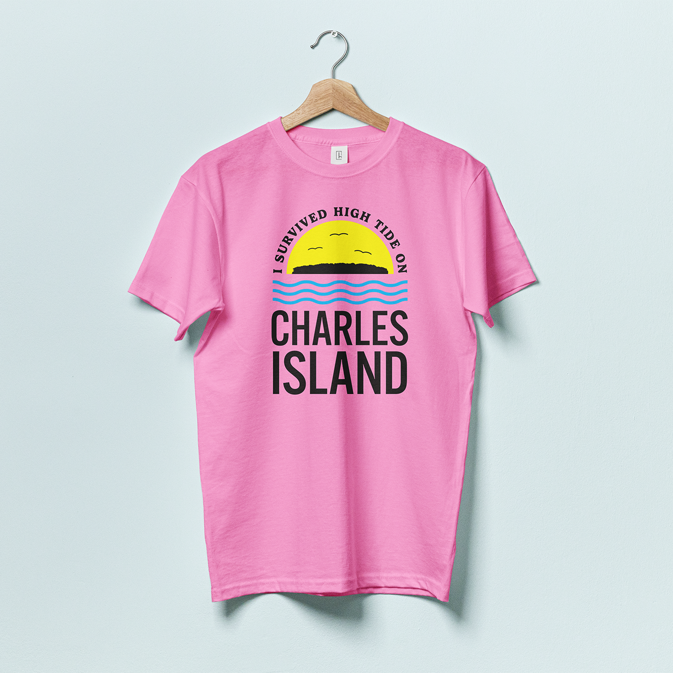 A pink t-shirt with a silhouette of Charles Island on top of a yellow semi circle, with blue wavy lines beneath and the words "I survived high tide on Charles Island"