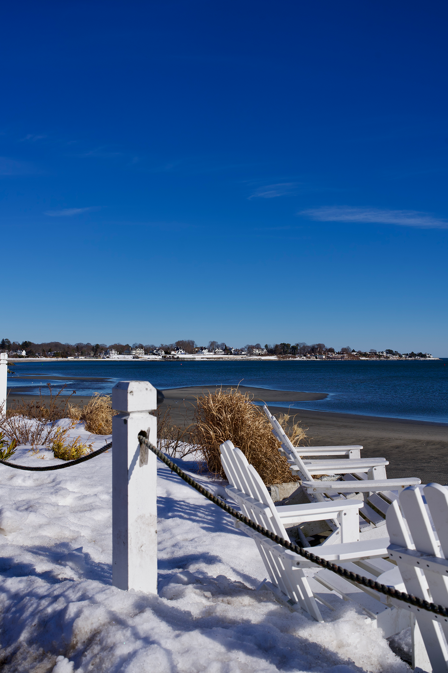 a snowy beach with Adirondack chairs and a view of the water