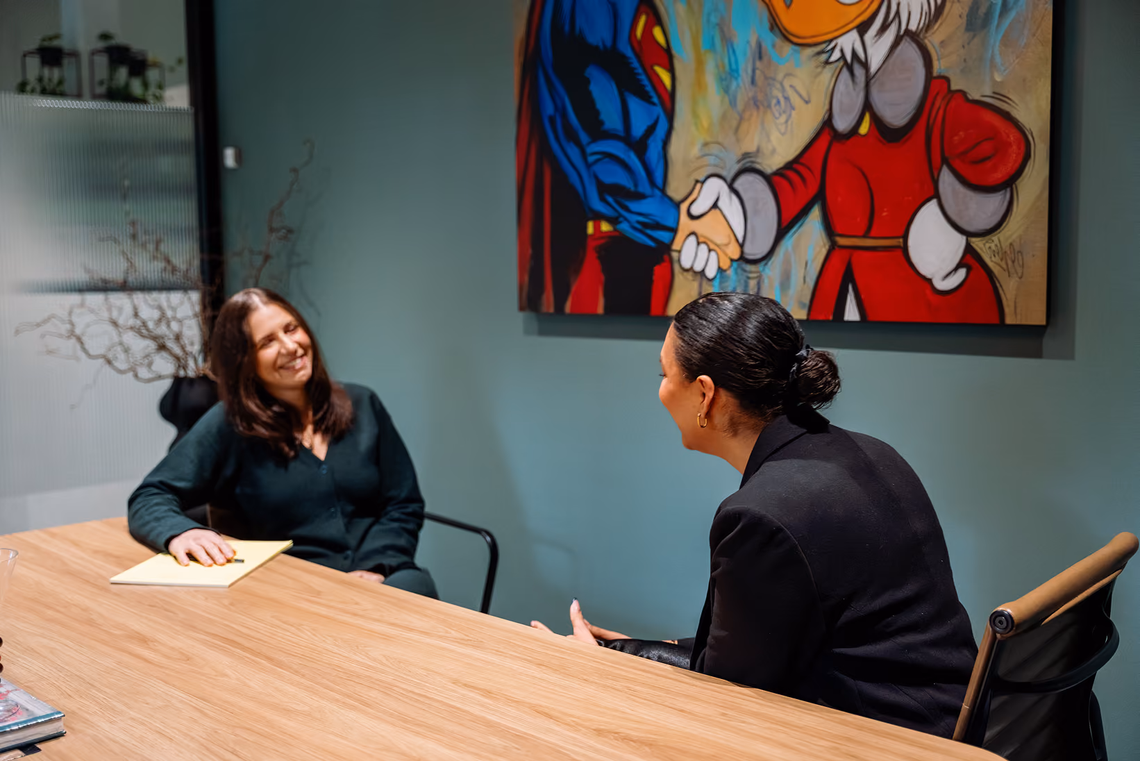 Two women sitting at a table in an office, engaged in a friendly conversation with artwork behind them.