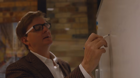 Man wearing glasses writing on a whiteboard with a marker in an indoor setting.