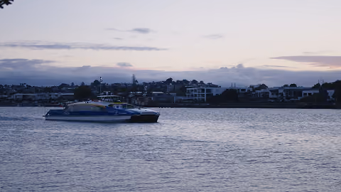 A catamaran boat floating on calm water near a coastal town at dusk.
