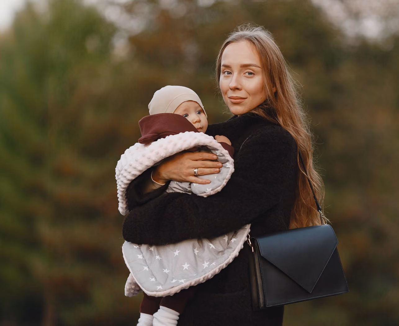 Woman wearing black coat holds baby wrapped in star-patterned blanket outdoors.