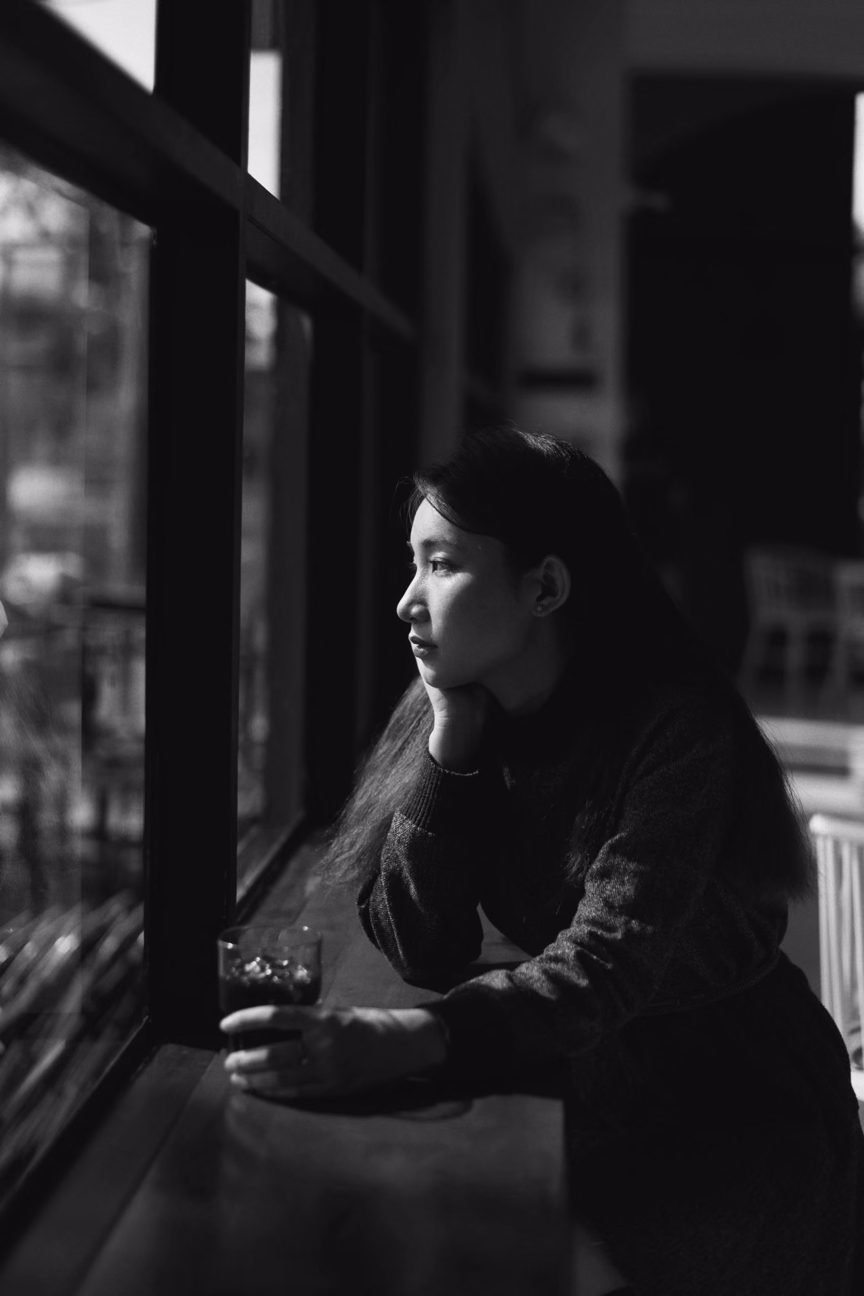Woman with long hair holding a drink and gazing pensively out a window in black and white photo.