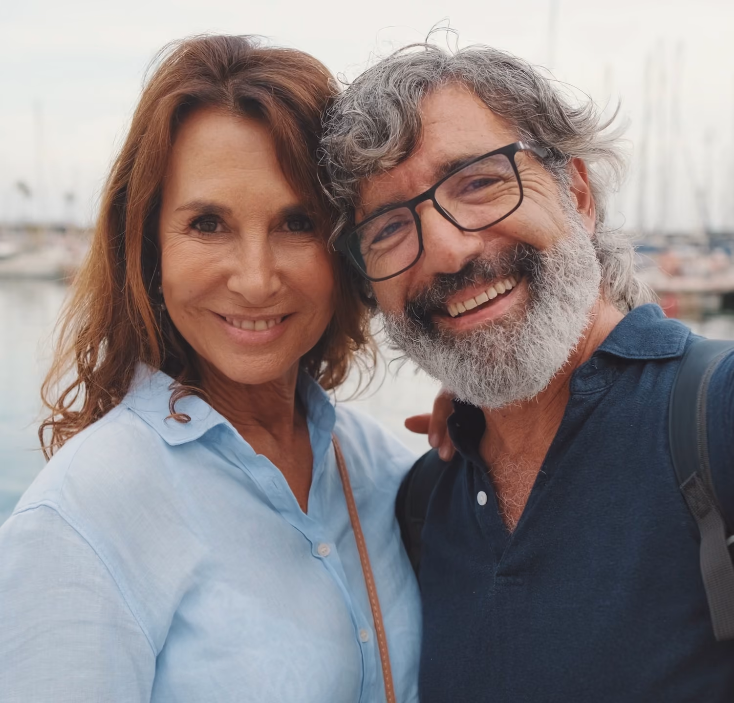Smiling middle-aged couple standing close by a marina with boats in the background.