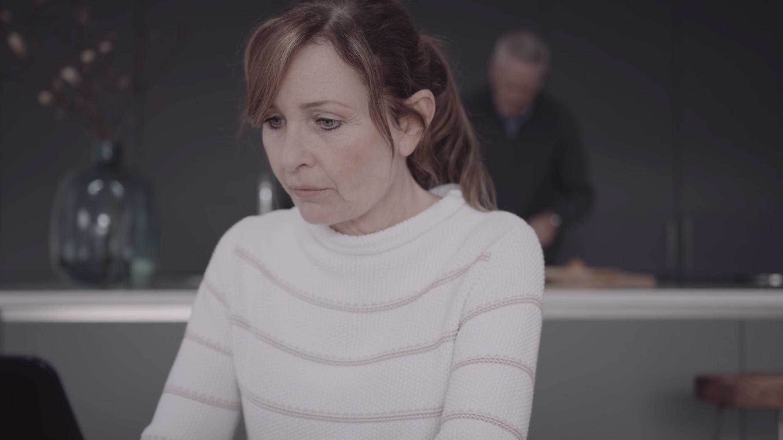 A woman in a white sweater looking pensive with a man blurred in the background in a kitchen.