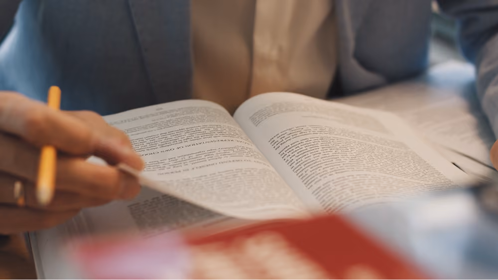 Person in a blazer turning pages of an open book with a pencil in hand.