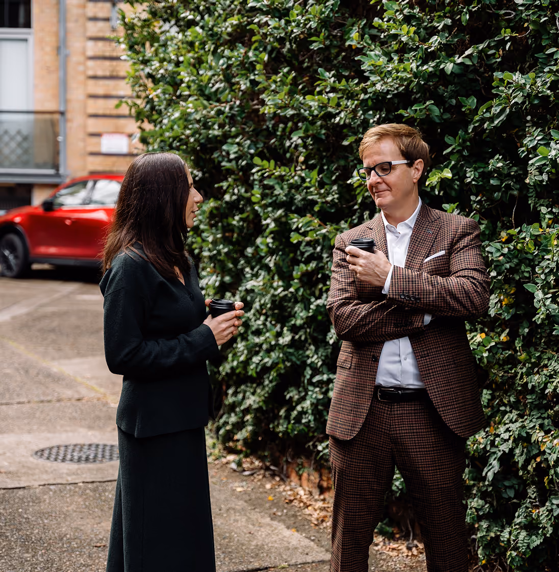Two people holding coffee cups engaged in conversation by a green leafy hedge outdoors.