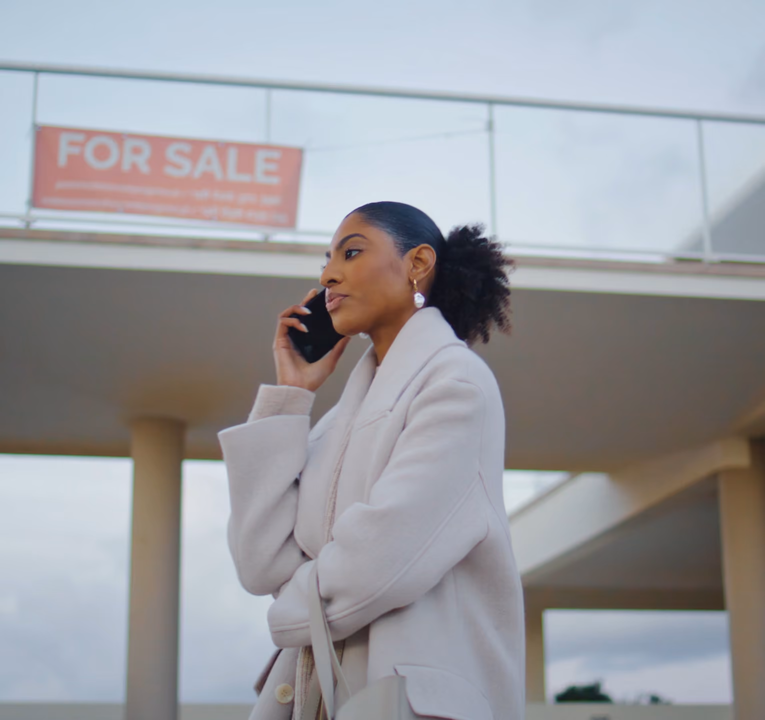 Woman in beige coat talking on phone outside a building with a 'For Sale' sign.