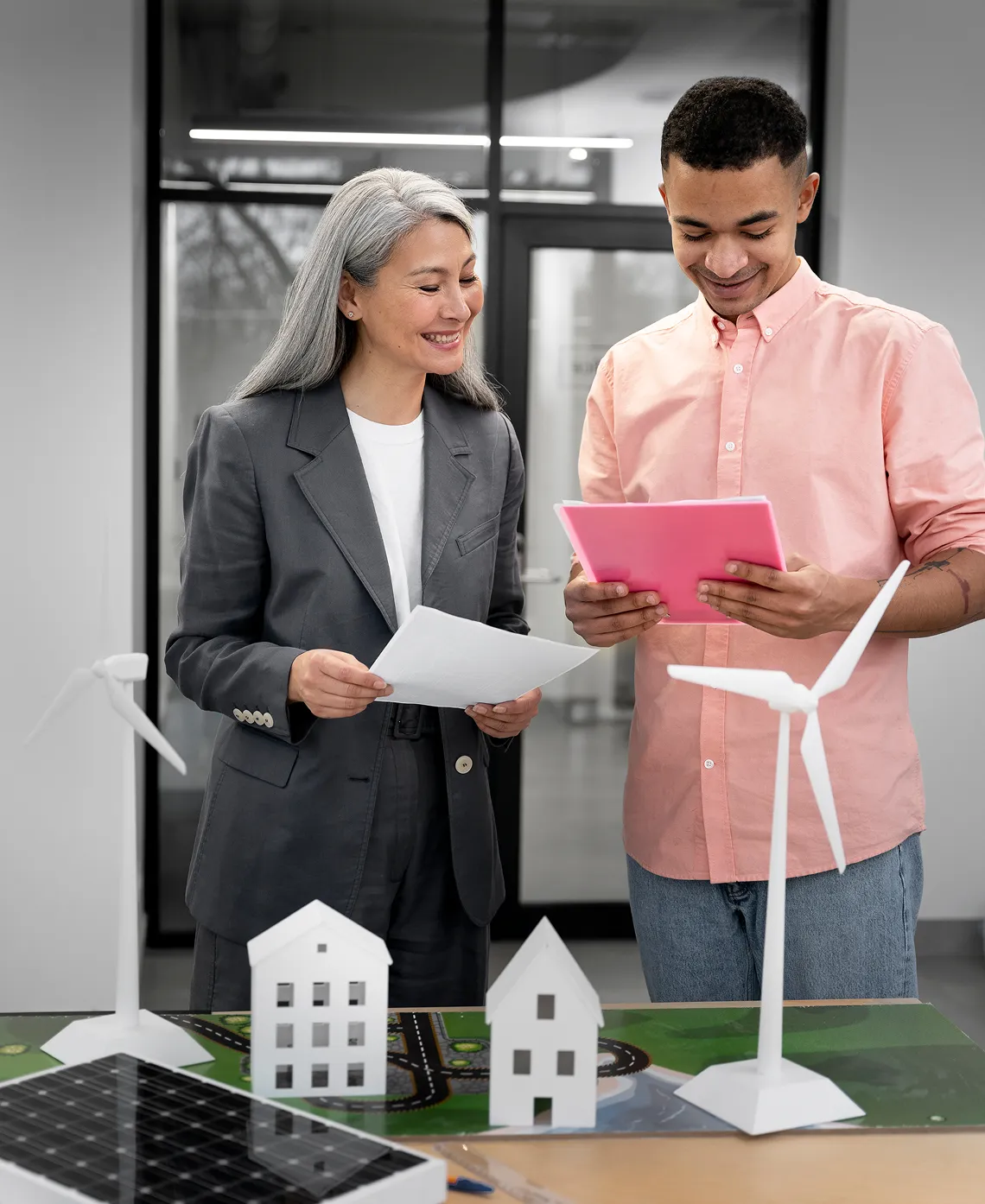 Two people reviewing documents near a table with miniature models of wind turbines, houses, and solar panels.