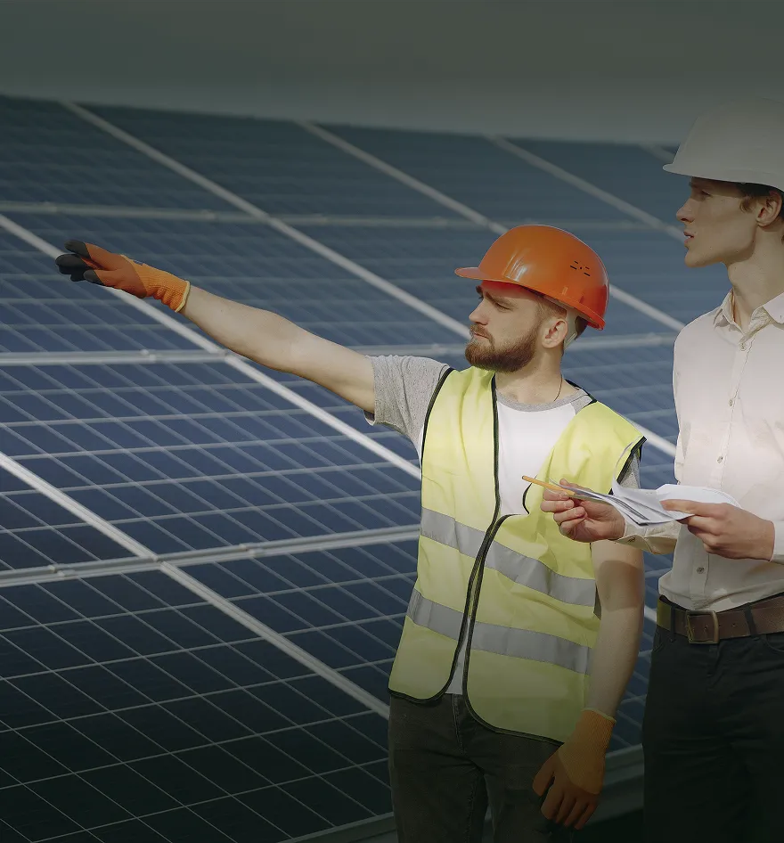 Two men wearing hard hats and safety gear stand by solar panels, one points ahead while the other holds papers and a pencil.