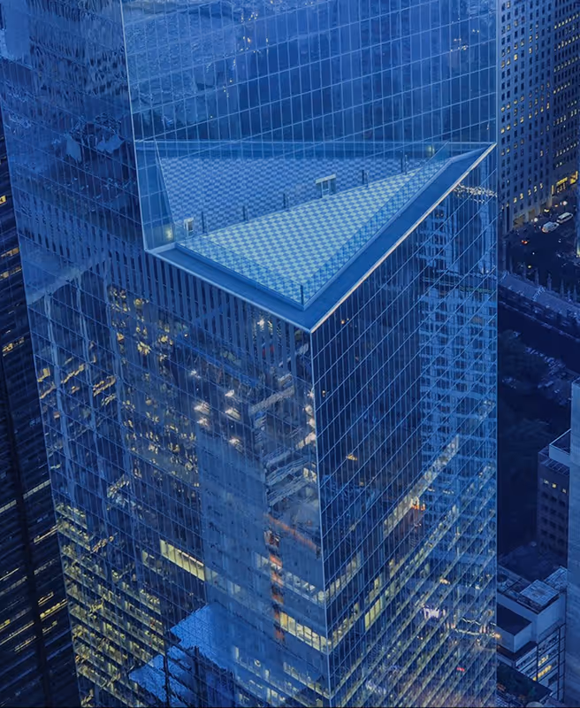 Aerial view of a modern glass skyscraper with a triangular rooftop section reflecting city lights at dusk.