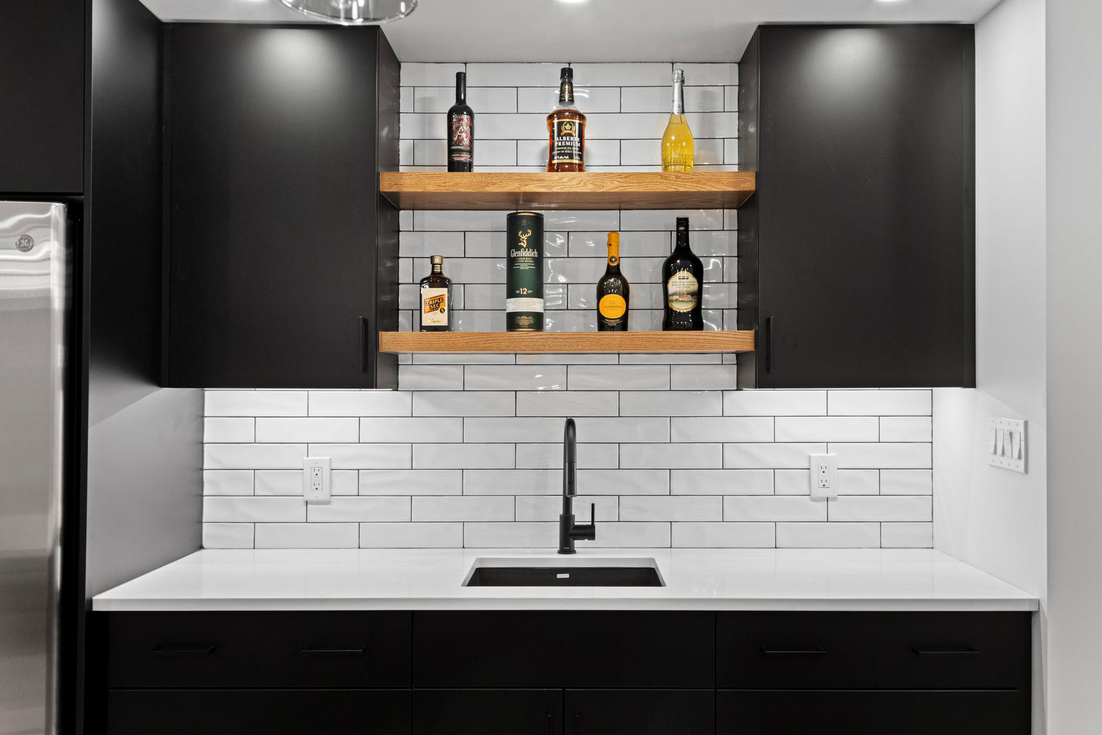 Modern kitchen bar area with black cabinets, white subway tile backsplash, and liquor bottles on wooden shelves.