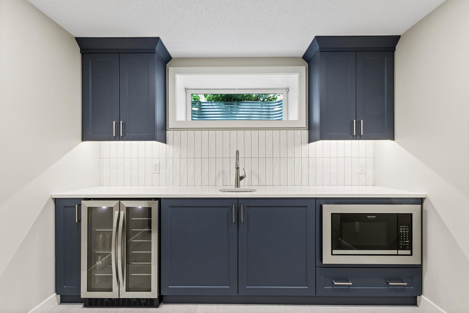 Modern kitchenette with navy cabinets, a sink under a small window, a wine fridge, and a built-in microwave.