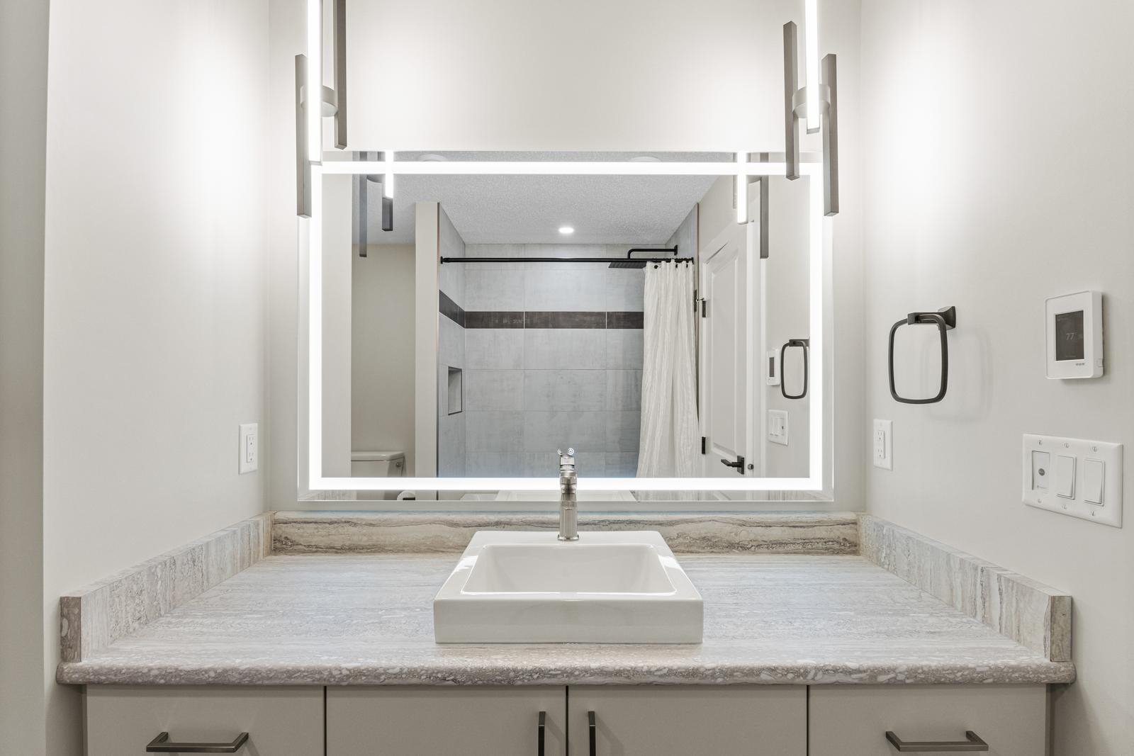 Modern bathroom vanity with illuminated mirror, square sink, and marble countertop in neutral tones.