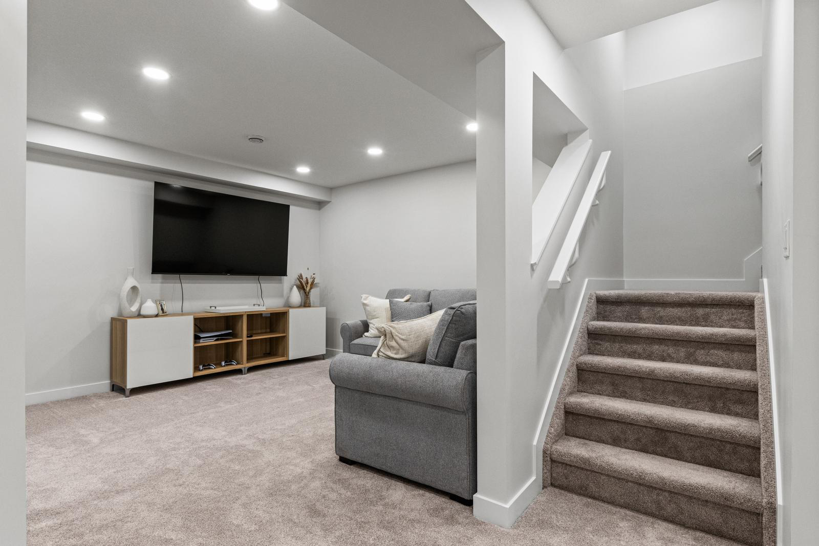 Modern basement living area with gray couch, flat-screen TV on wall, and carpeted stairs.