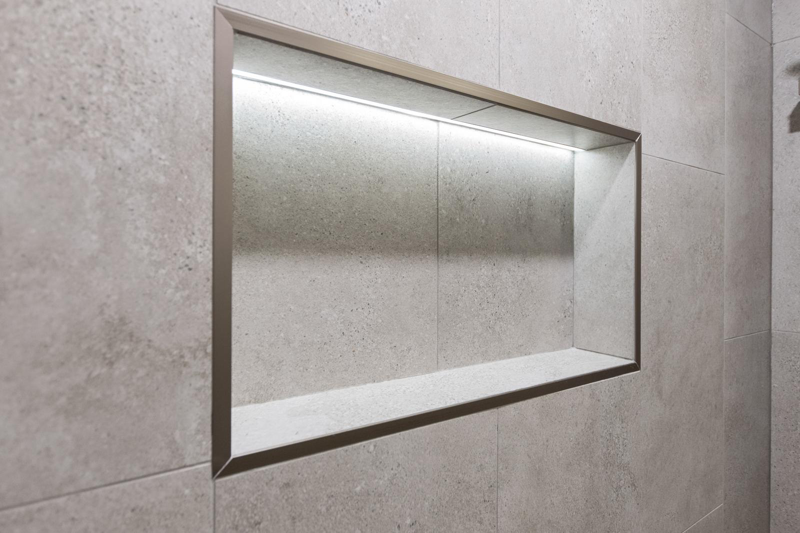 Rectangular recessed niche with LED strip lighting framed in matte metal in a tiled wall.