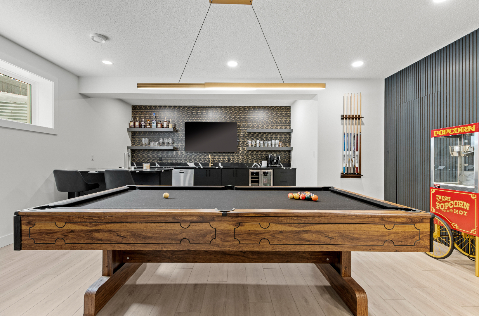 Modern game room with a wooden pool table, black felt, bar area, TV, and a red popcorn machine.