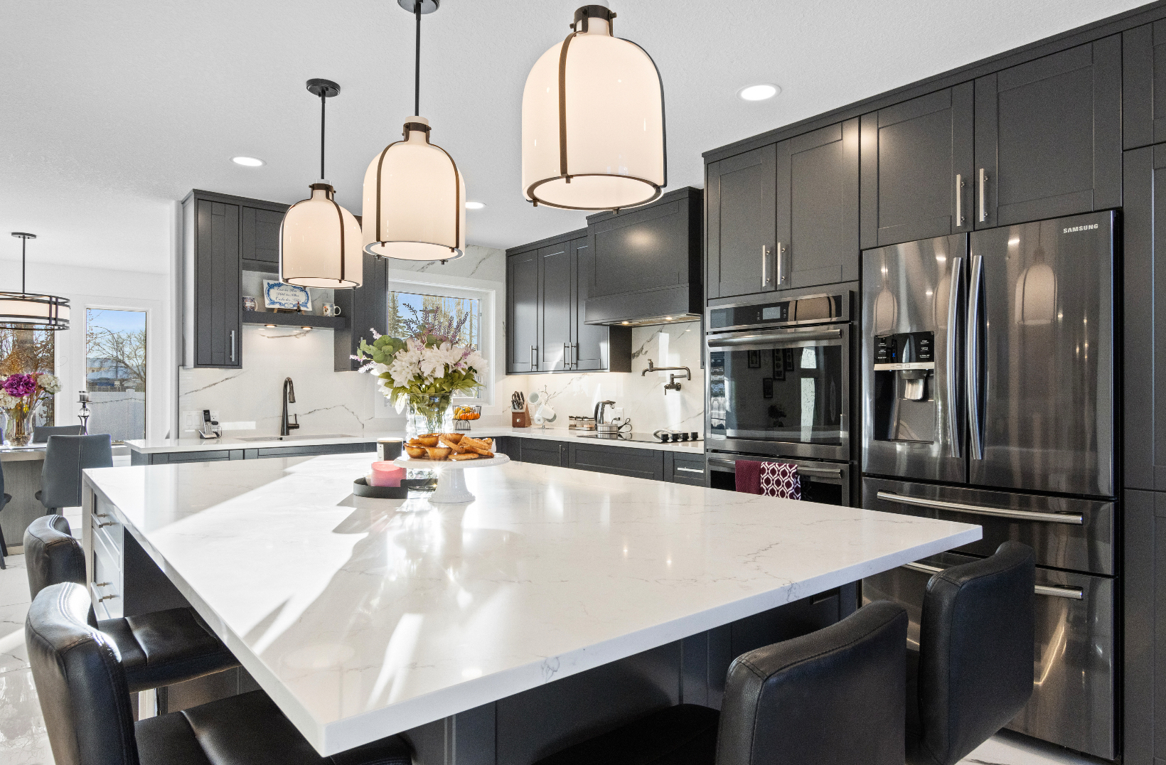 Modern kitchen with large white island, black leather stools, dark cabinets, and stainless steel appliances.