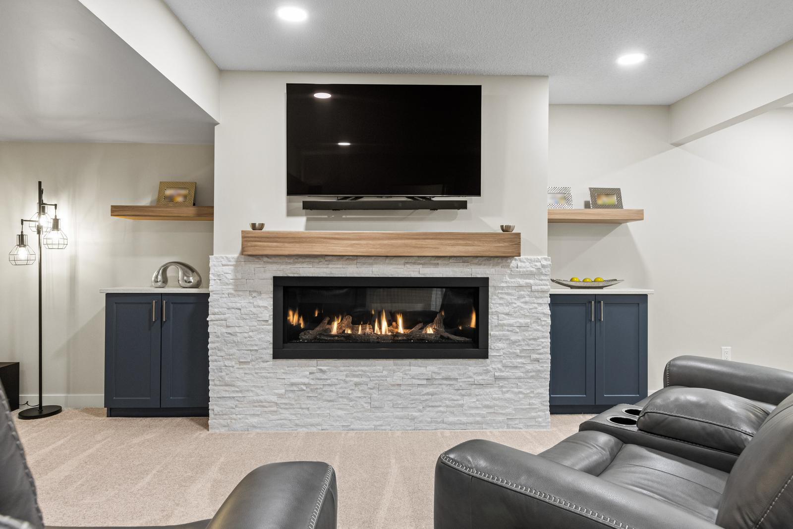 Modern living room with a gas fireplace, TV above, floating shelves, navy cabinets, and leather recliners.