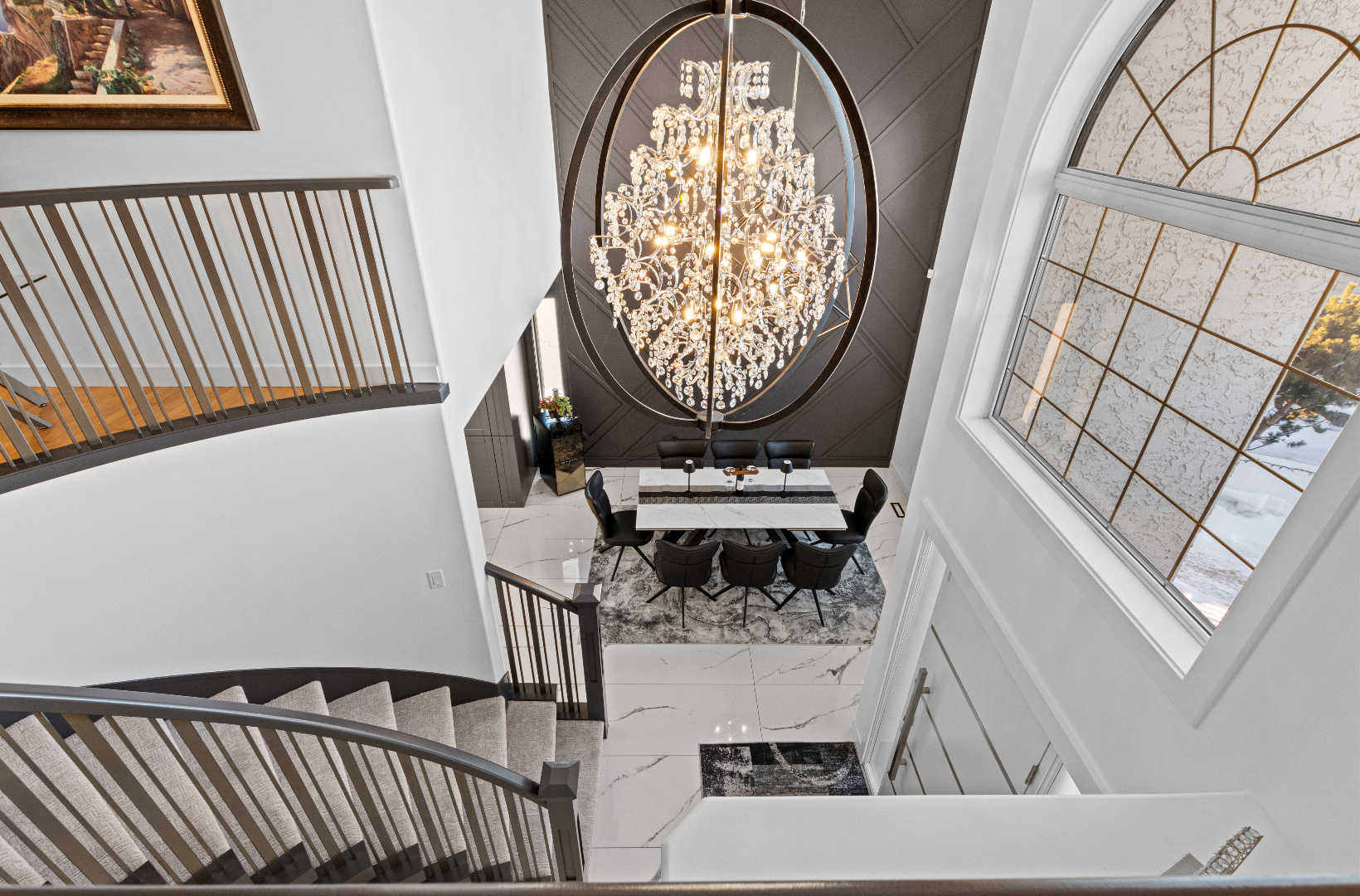 Modern dining area below a large crystal chandelier and curved staircase with metal railings.