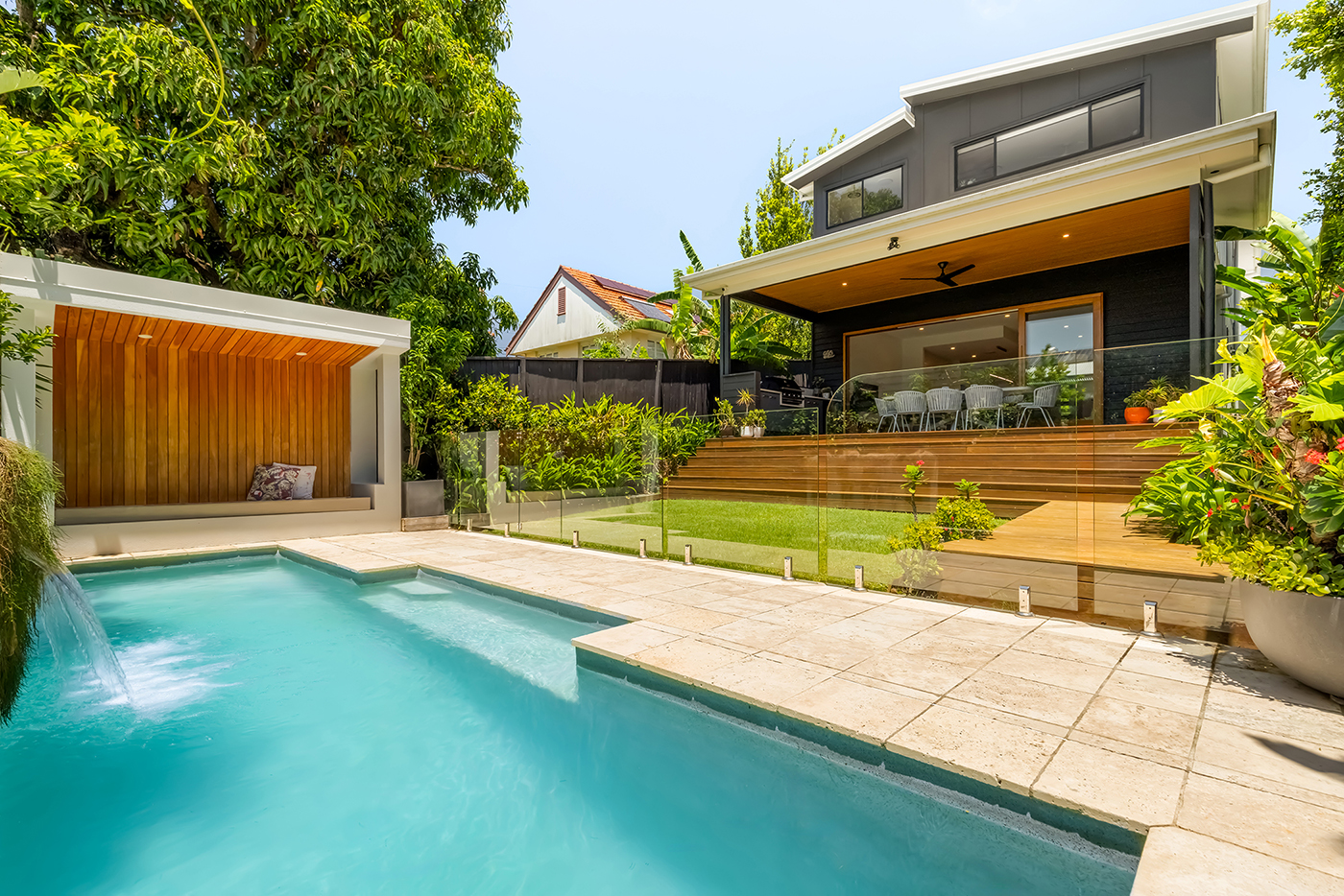 Modern backyard with a blue swimming pool, a wooden seating nook, glass fence, and a covered deck with outdoor dining set.