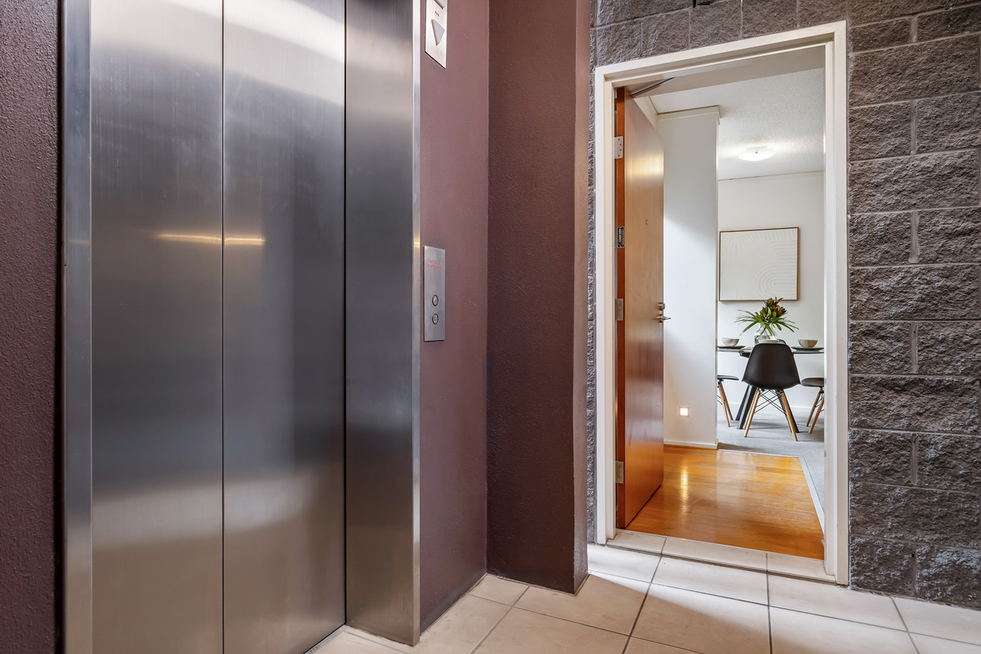 Closed stainless steel elevator doors next to an open doorway showing a modern dining area with black chairs and a framed artwork.