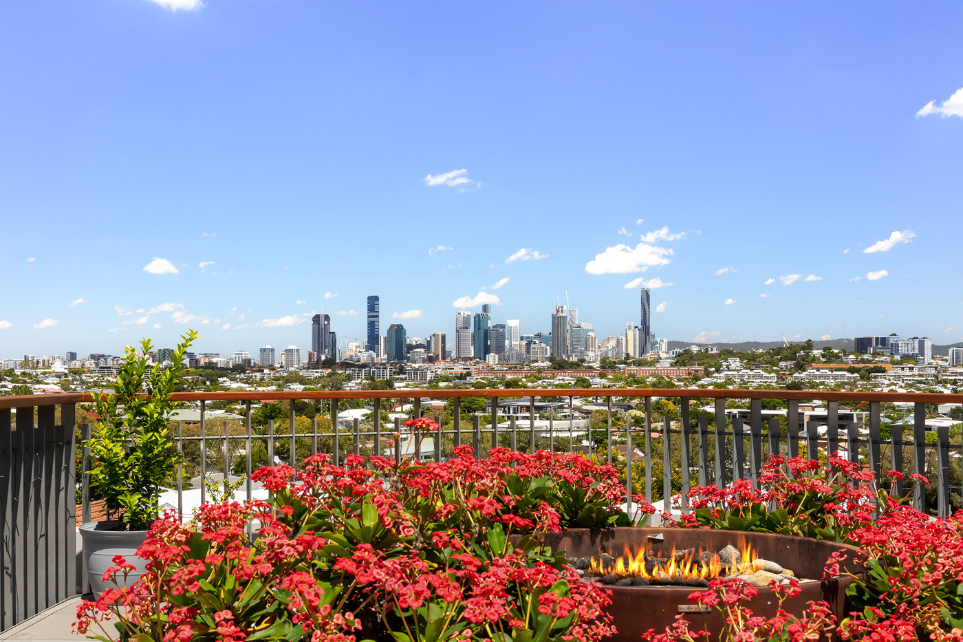 City skyline viewed from a balcony with a fire pit surrounded by red flowers and potted plants under a clear blue sky.