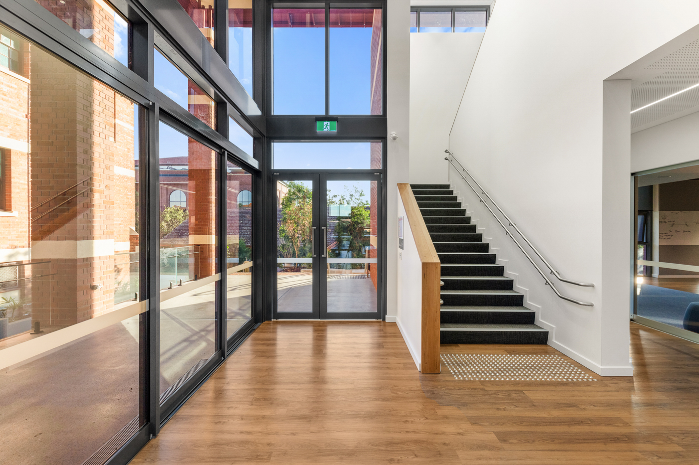 Bright modern hallway with wooden floor, black-framed glass doors and windows, and a staircase with black carpet steps and metal handrails.