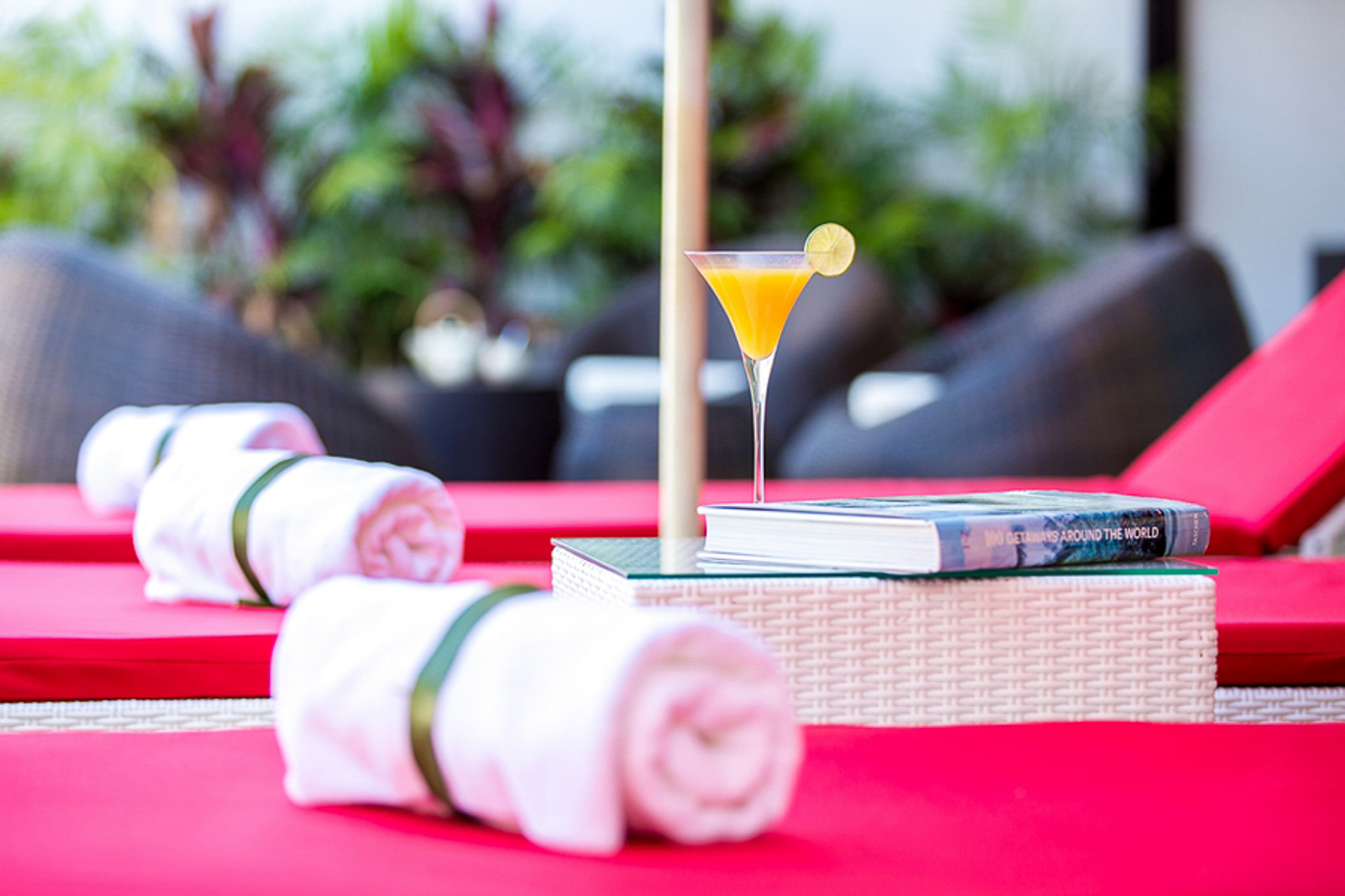 Rolled white towels on red cushioned lounge chairs with a cocktail glass garnished with lime and a book on a wicker table by a poolside.
