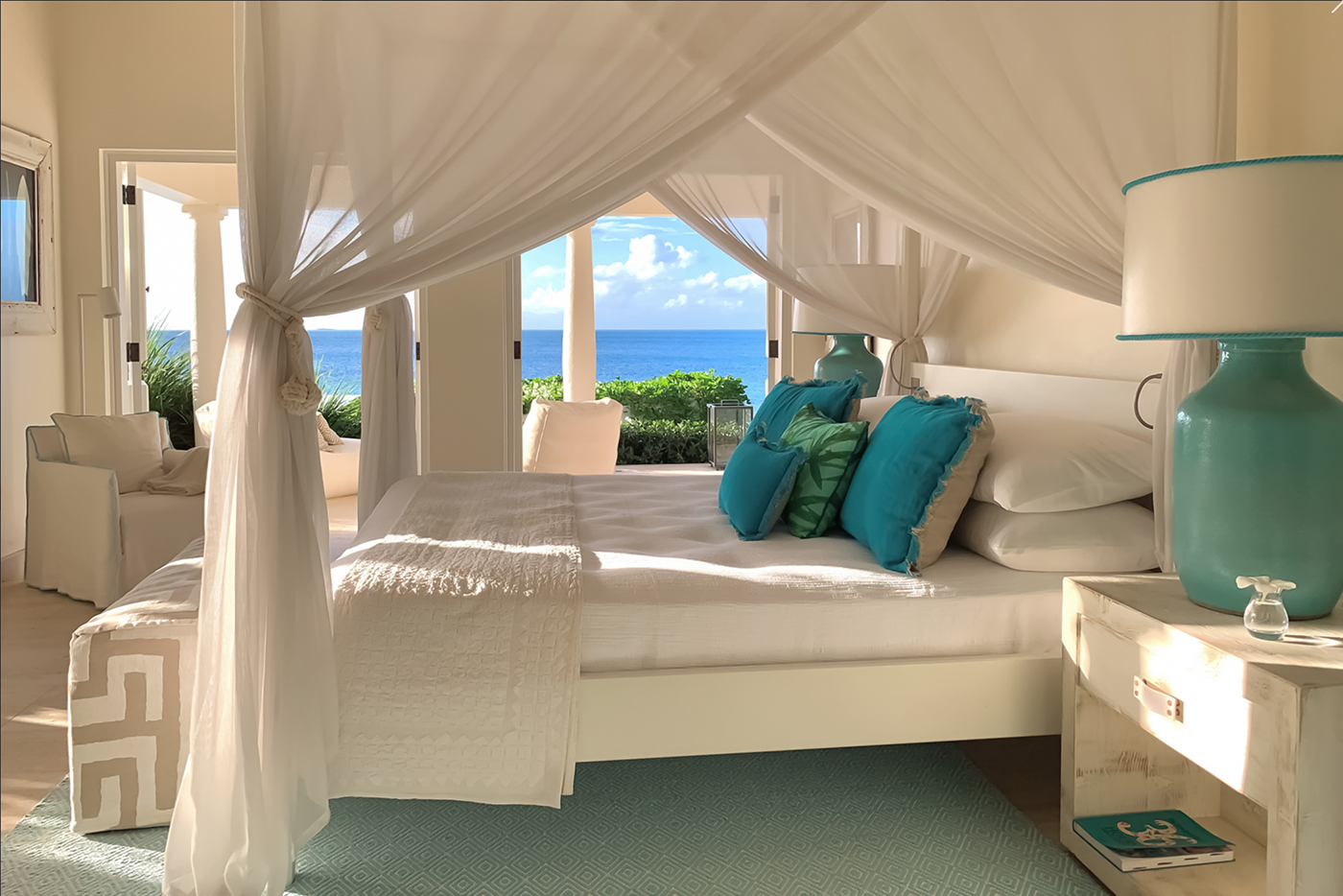 Airy bedroom with white canopy bed, teal cushions, and an ocean view through open doors.