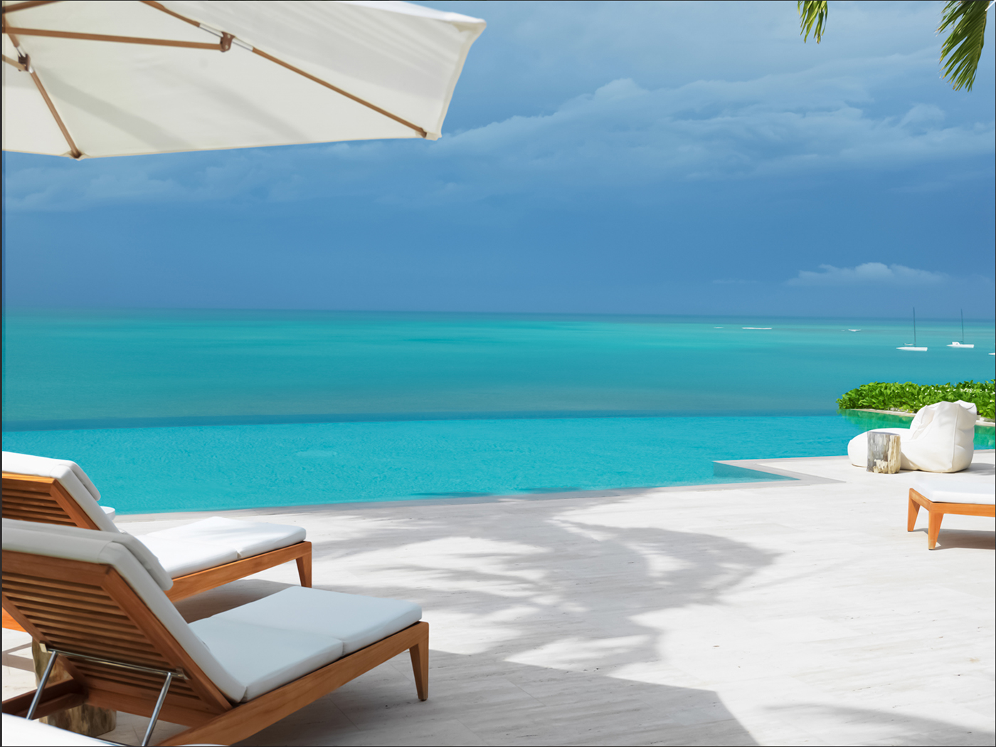 Infinity pool overlooking turquoise ocean with white lounge chairs and a large umbrella on a sunny patio.