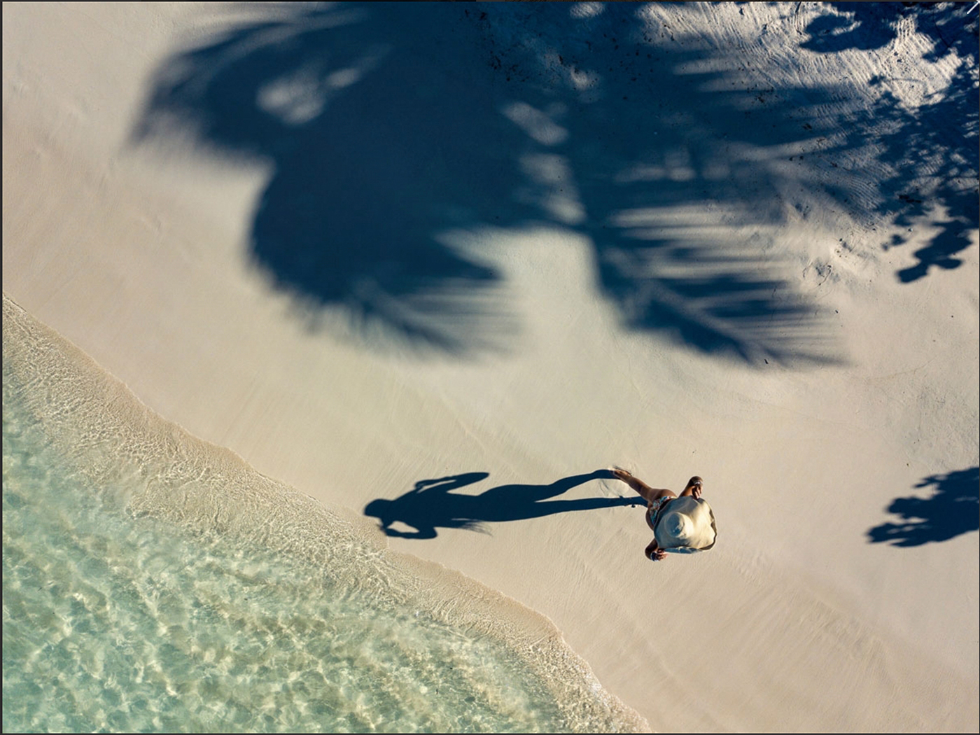 A person walking barefoot on white sand near clear ocean water with large palm tree shadows on the beach.