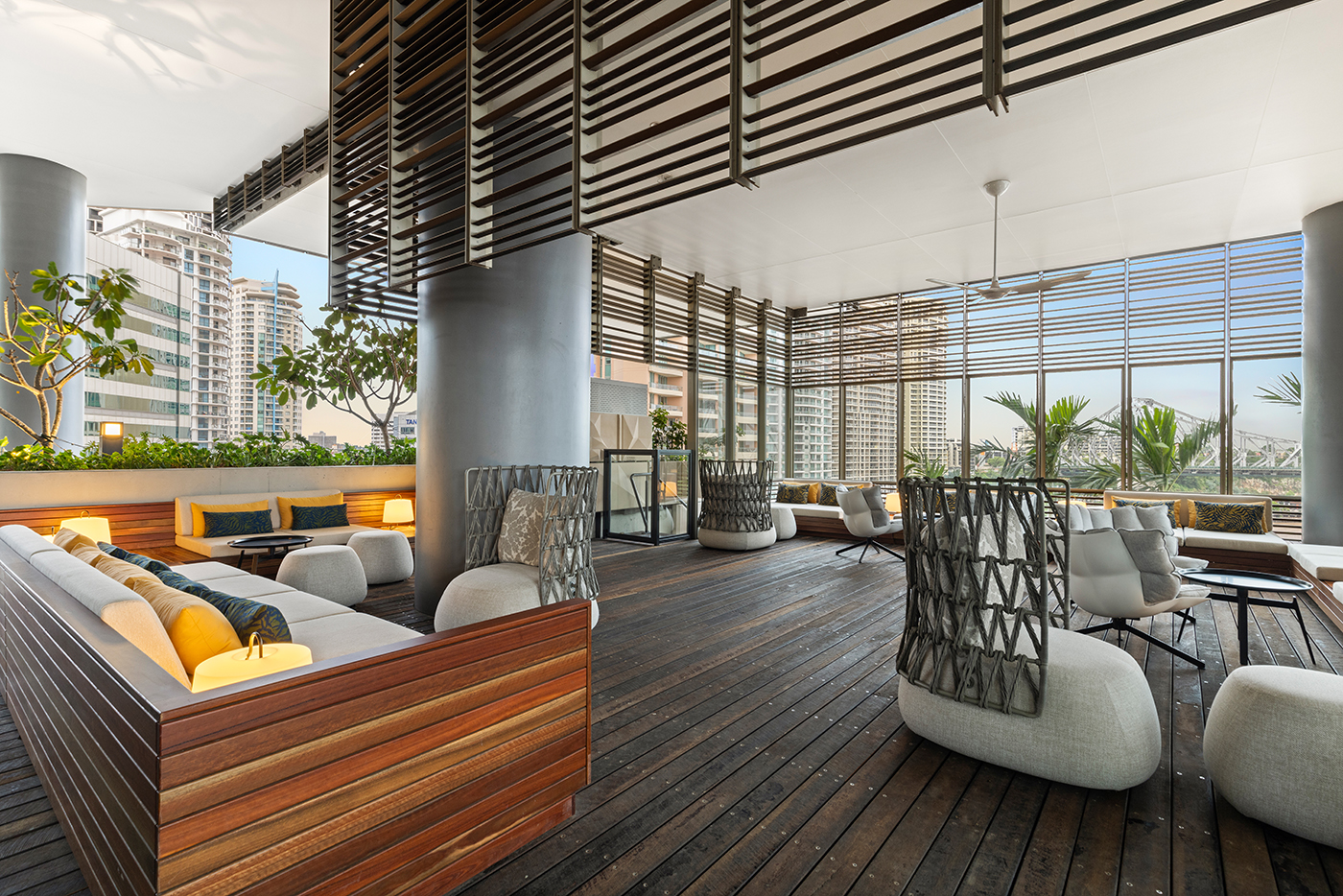 Modern outdoor lounge area with wooden flooring, cushioned seating, potted plants, and cityscape views through large slatted windows.