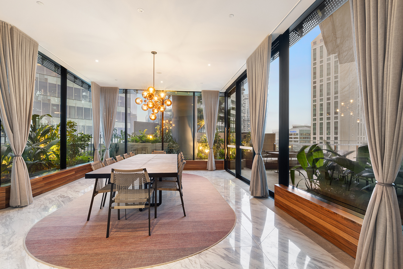 Modern dining room with a long table, woven chairs, large windows with beige curtains, plants outside, and a spherical pendant light.
