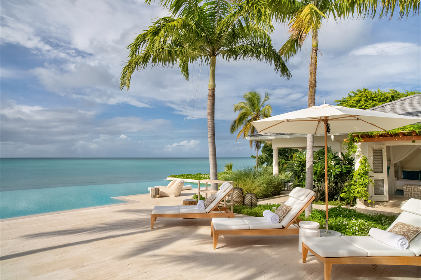 Luxury poolside with white cushioned sun loungers, umbrellas, palm trees, and ocean view under a partly cloudy sky.
