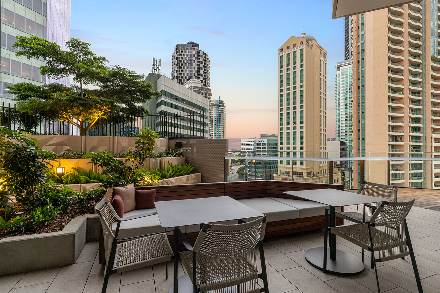 Modern outdoor terrace with tables, woven chairs, cushioned bench seating, plants, and city buildings in the background at sunset.