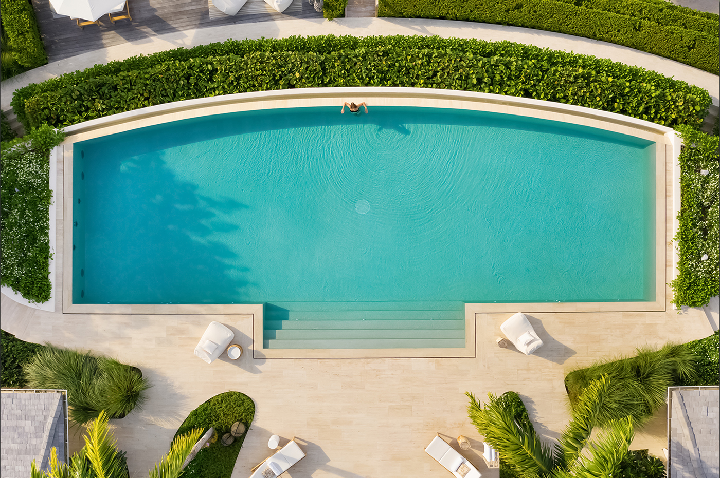 Aerial view of a person relaxing alone in a turquoise rectangular swimming pool surrounded by green bushes and lounge chairs.