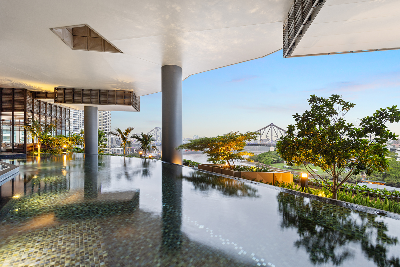 Modern outdoor infinity pool with mosaic tiles overlooking a city bridge and river during sunset.